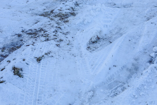 A snow-covered road with deep tire tracks illustrates challenging winter driving conditions. Frosty snow cover is mixed with mud and frozen ground under cold light. The textured surface captures the a