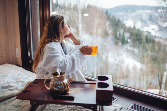 Drinking hot tea enjoying winter landscape in mountains. Woman wears bathrobe relaxing by big window of cabin house on holiday