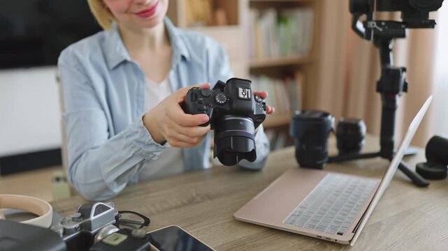 A smiling caucasian woman holds a digital camera while surrounded by professional photography equipment and a laptop on a wooden desk
