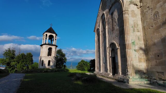 Historical nikortsminda cathedral revealing its intricate stone facade and a separate bell tower under a bright blue sky. A significant monument of medieval georgian architecture in the racha region