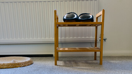 Black slippers with white fleece lining on a two-tier wooden shoe rack placed on carpeted floor near radiator. © Kos6ka