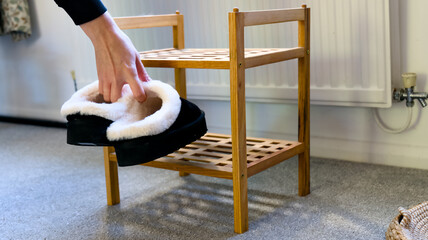 Person holding black house slippers with soft fleece lining near a wooden shoe stand indoors. © Kos6ka