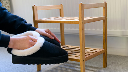 Hands holding cozy black house slippers with plush white lining next to a small wooden shoe rack on grey carpet. © Kos6ka