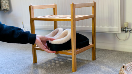 Black winter slippers with white fleece lining placed on a small wooden shoe rack on a grey carpeted floor. © Kos6ka