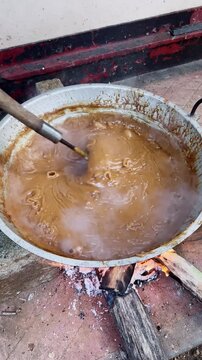sweet dodol candy in large metal pot over wood fire, bubbling sticky brown mixture stirred outdoor cooking scene, traditional asian dessert preparation