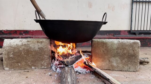 big black wok cooking over open wood fire on concrete blocks, burning logs and bright orange flames heating large metal pan, traditional outdoor culinary preparation in slow motion
