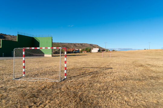 An open rural recreation ground features two small red and white goalposts on dry grass beside a tall green fronton wall used for pelota games. The unmarked field, distant houses and rolling hills ill