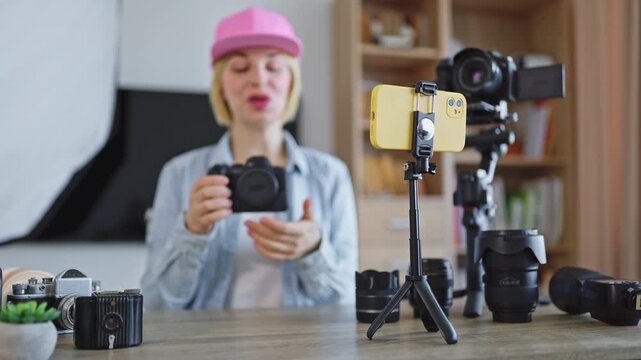 A caucasian woman in a pink cap and denim shirt is filming herself with a DSLR camera and smartphone on a tripod, surrounded by lenses and equipment