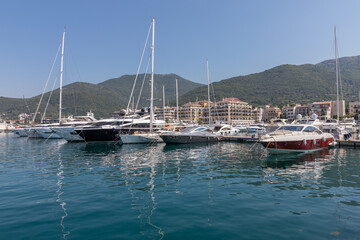 Obraz premium Tivat, Montenegro - August 11, 2025: Moored luxury yachts and motorboats rest on calm blue sea waters along coastal resort pier in Tivat, surrounded by mountain slopes and modern harbor architecture