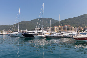 Obraz premium Tivat, Montenegro - August 11, 2025: Luxury yachts and motorboats rest in calm harbor waters of Tivat, Montenegro, featuring coastal architecture and lush green hills under bright summer sky