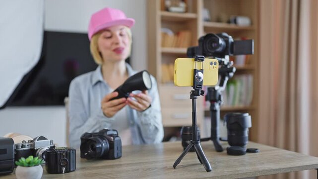 A caucasian woman in a pink cap reviews camera equipment for her online channel, surrounded by various cameras and lenses