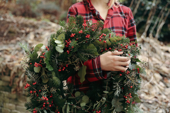 Woman in red pyjamas Holding Large Handmade Christmas Wreath with Fresh Fir Branches, Eucalyptus and Berries Outdoors. Natural Evergreen Holiday Decoration. Festive Winter Holiday, Blurred background