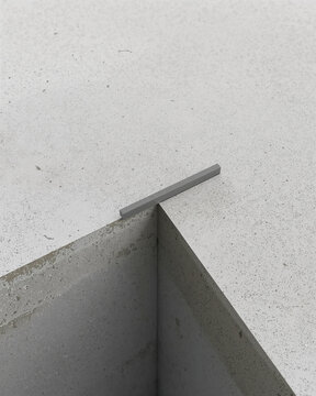A small gray rectangular bar placed on the edge of a concrete block, creating a sharp geometric corner with a deep, dark, empty vertical space below