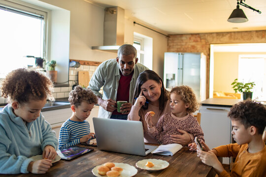 Parents multitasking at laptop with children using devices in kitchen