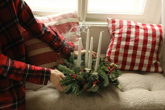 Kid in checkered pyjamas holding Decorative Traditional Christmas Advent Wreath with White Smoking Candles, Fir Branches and Red Berries in Cozy Festive Home Interior. Winter Holiday Preparation