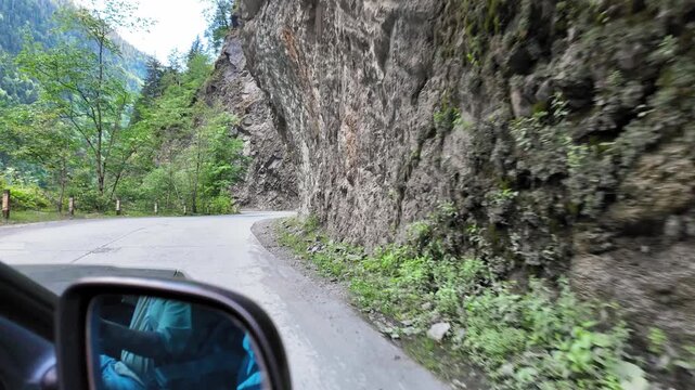 Personal point of view from a vehicle driving along a narrow and hazardous mountain pass, with a steep rock wall on one side and a lush green valley on the other, showing signs of recent rockfalls