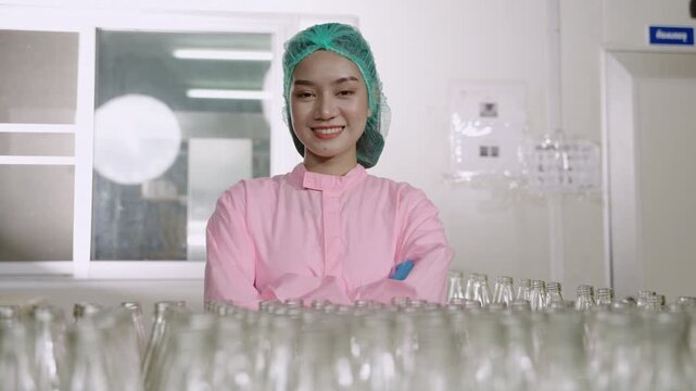 Happy beautiful Asian woman technician inspector wearing a uniform of factory standing smiling with arms crossed while looking at camera on beverage bottle production line in manufacturing industry. 