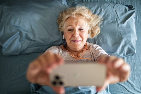 Senior woman in bed taking smartphone selfie with earbuds