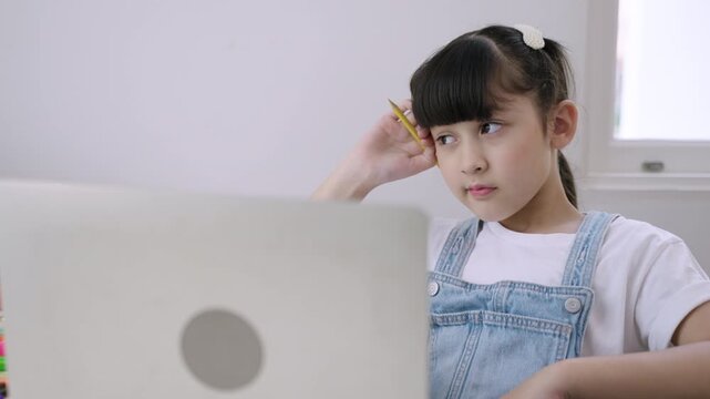 Cute Asian girl student sitting at a desk using a laptop computer for online learning and thinking of an answer via webcam with a remote teacher in homeschooling. New normal of the education concept. 
