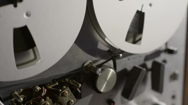 Vintage reel-to-reel tape recorder operates mechanical heads in an audio studio close-up. Exposed pinch rollers, magnetic heads, and tape path mechanisms enrich technical documentaries.