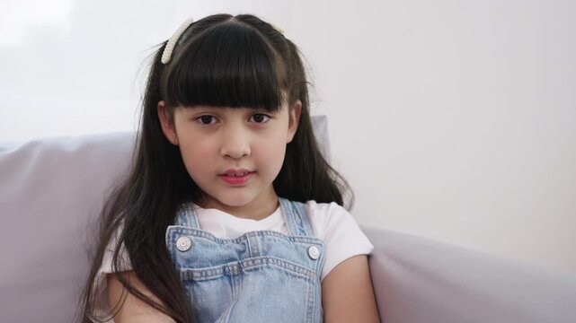 A happy, cute Asian girl is sitting on a sofa in her living room at home and posing for the camera. Confident, adorable model child girl looking at the camera and gesturing on the couch in the studio.