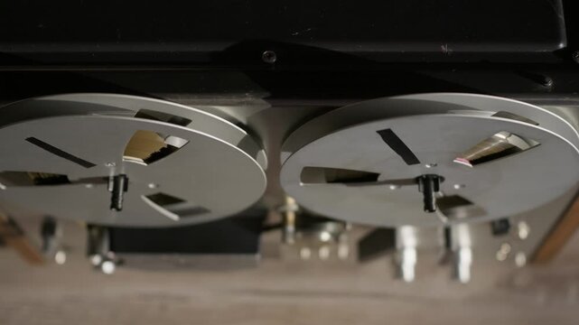 Vintage reel-to-reel tape deck steadily spins magnetic spools inside an analog audio studio. This overhead perspective highlights the dual rotating metal reels and upper deck layout.