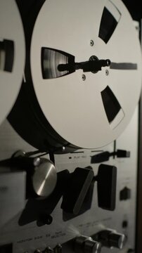 A vintage reel-to-reel tape deck spins steadily inside a dark broadcast studio setting. Dramatic side lighting highlights the retro audio gear for an authentic broadcasting mood.