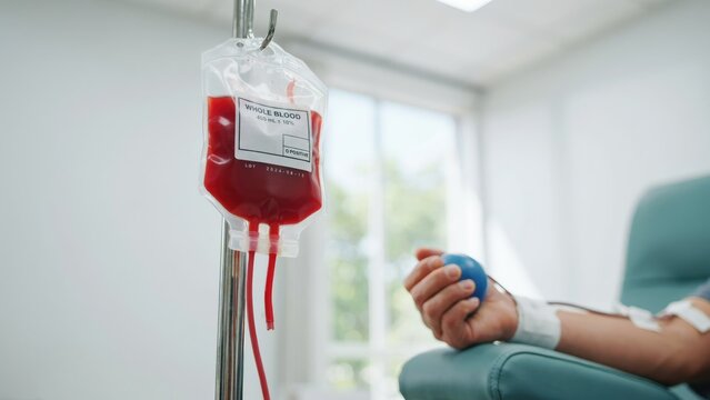 Whole donation bag hanging on a metal stand, with a donor squeezing a blue stress ball in a medical clinic setting
