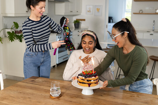 Diverse female friends presenting layered cake with topper in kitchen, woman wearing tiara