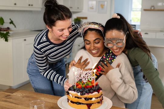 Diverse female friends celebrating birthday at kitchen table with layered berry cake and candles