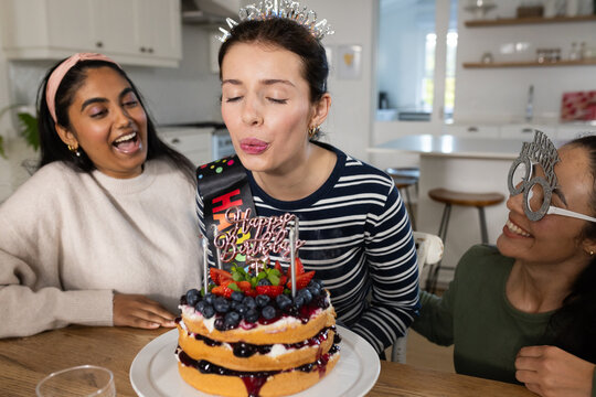 Female friends center wearing tiara and sash gathering around kitchen table blowing candles on cake