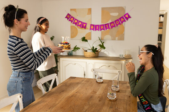 Diverse female friends bringing birthday cake with lit candles toward seated woman at dining table