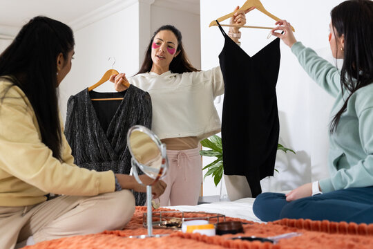 Diverse female friends comparing dresses on bed in bright bedroom, holding wooden hangers, mirrors