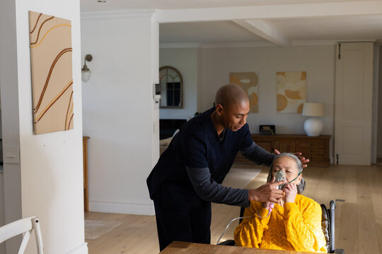 African American carer in scrubs leaning, placing oxygen mask tubing on senior Asian woman at table