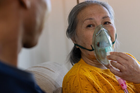 Senior Asian woman sitting on sofa wearing yellow sweater holding oxygen mask with pink tubing