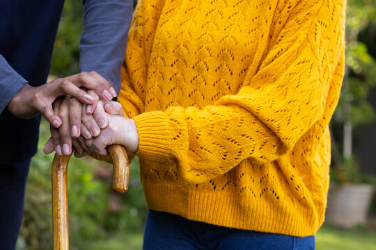 Diverse man and Senior Asian woman placing hands on wooden cane in garden, wearing yellow sweater