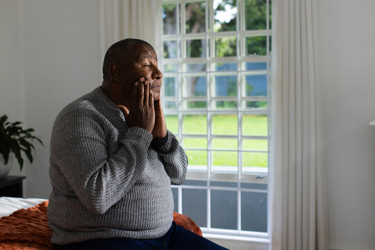 Senior African American man sitting on bed pressing hands to cheeks near window with rust throw