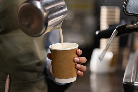 Green-aproned Asian female barista pouring milk into paper cup at coffee counter with steel pitcher