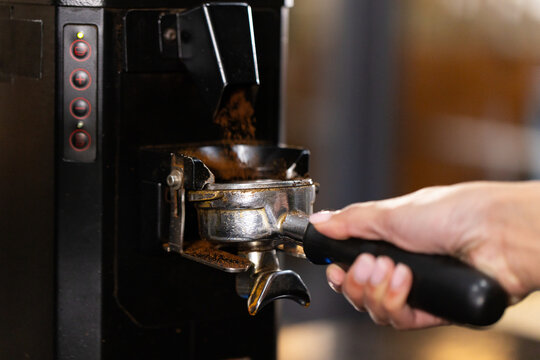 Asian woman holding portafilter with coffee grounds falling from espresso grinder at cafe