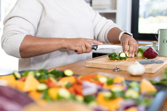 Mature African American woman wearing cream sweater slicing zucchini with chef knife on countertop
