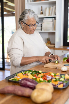 Senior African woman slicing zucchini in kitchen at island, wearing sweater with knife, baking tray