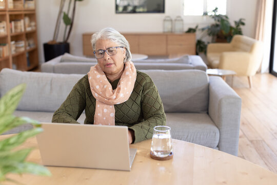 Senior female sitting at table in living room using laptop, wearing green sweater and pink scarf