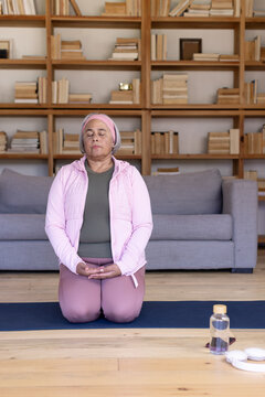 Senior African American woman kneeling, meditating on mat at home with pink headband, water bottle
