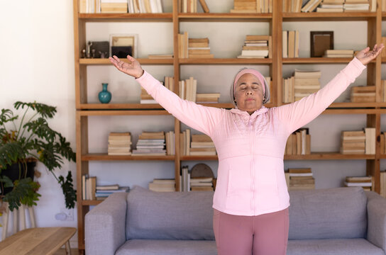 Senior African American woman stretching, breathing in living room by bookcase, wearing pink top