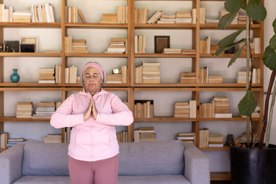 Senior African American woman wearing pink hoodie standing in library by bookshelf pressing palms