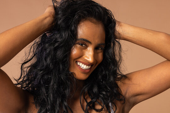 Female model posing, running hands through curly hair in beige studio wearing thin-strap top