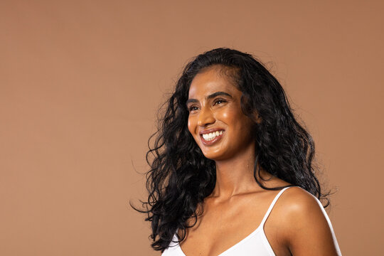 Adult Indian woman smiling and wearing white spaghetti-strap camisole in studio with tan backdrop