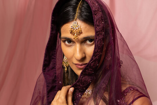 Indian woman holding maroon embroidered dupatta near chin in studio, showcasing gold maang tikka