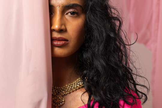 Indian woman peeking behind pale pink curtain in studio wearing bright pink top and gold necklace