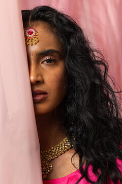 Woman posing while hiding face behind pale pink fabric panel in studio, wearing maang tikka